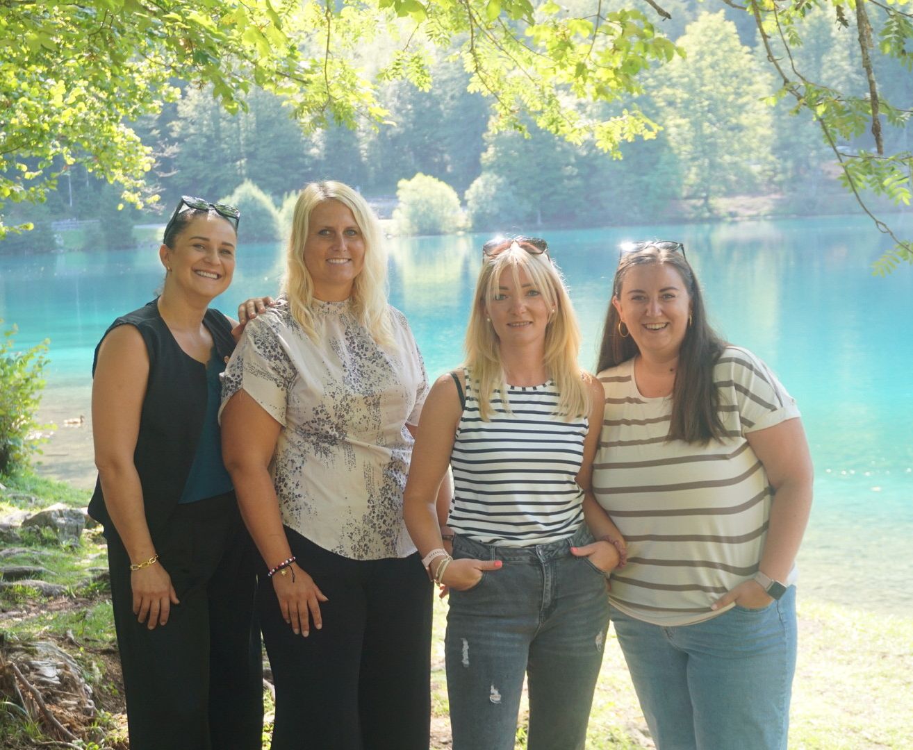Vier Frauen stehen zusammen vor einem See und posieren lächelnd für ein Foto. Sie tragen Freizeitkleidung und einige haben Sonnenbrillen auf dem Kopf. Der See ist von Bäumen umgeben und der Himmel ist hell.