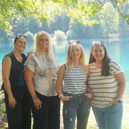 Vier Frauen stehen zusammen vor einem See und posieren lächelnd für ein Foto. Sie tragen Freizeitkleidung und einige haben Sonnenbrillen auf dem Kopf. Der See ist von Bäumen umgeben und der Himmel ist hell.