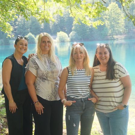 Vier Frauen stehen zusammen vor einem See und posieren lächelnd für ein Foto. Sie tragen Freizeitkleidung und einige haben Sonnenbrillen auf dem Kopf. Der See ist von Bäumen umgeben und der Himmel ist hell.