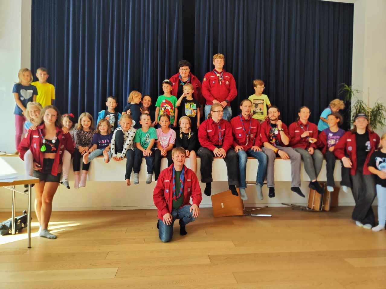 Eine Gruppe von Kindern und Erwachsenen, einige in roten Jacken, posiert für ein Foto auf einer Bühne mit blauem Vorhang im Hintergrund.