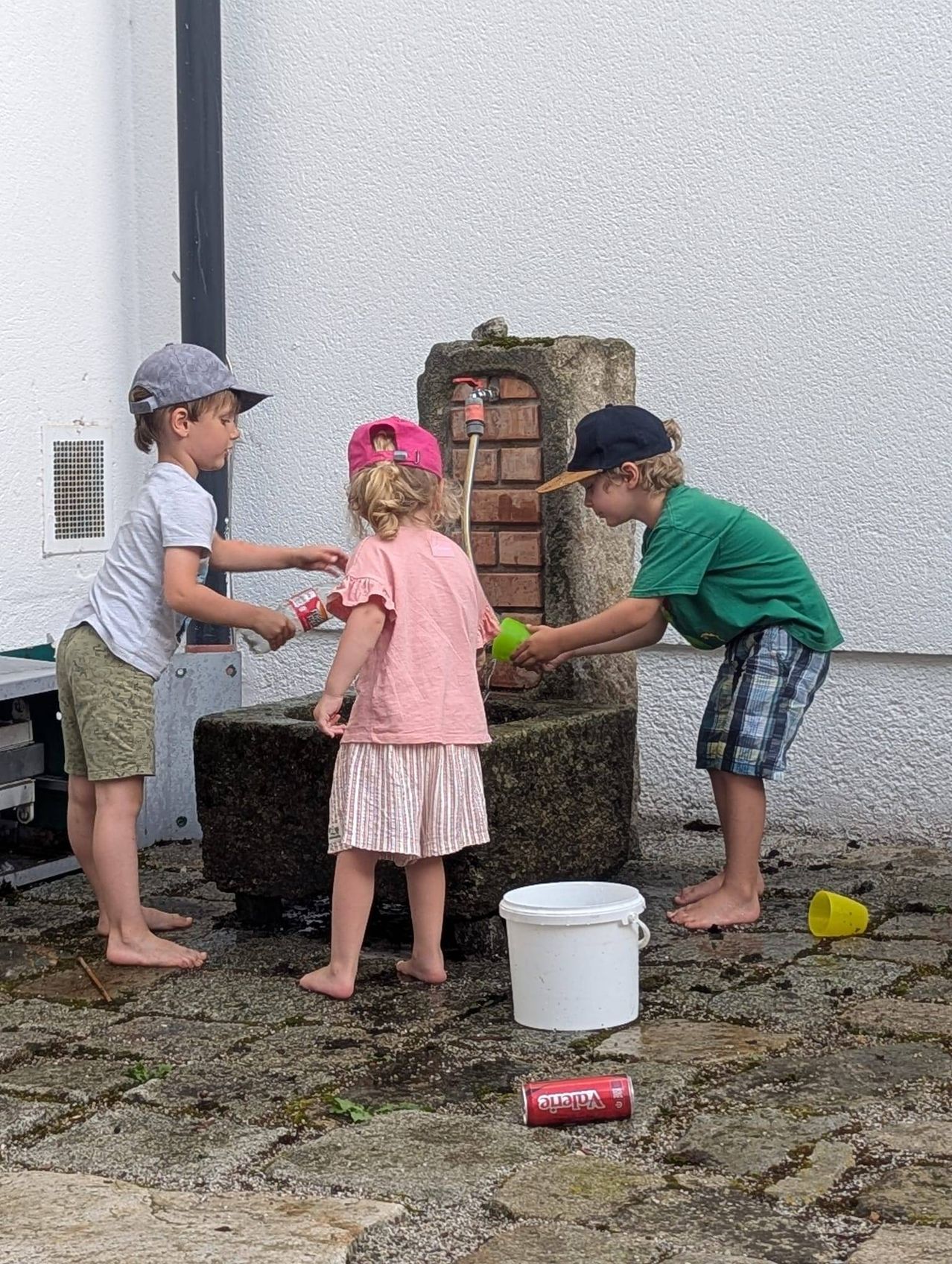 Drei Kinder, zwei Jungen und ein Mädchen, spielen in der Nähe eines Brunnens, einer mit einem roten Becher, einem anderen mit einem grünen Becher und das Mädchen hält eine Flasche. Sie sind barfuß und ein weißer Eimer ist in der Nähe.
