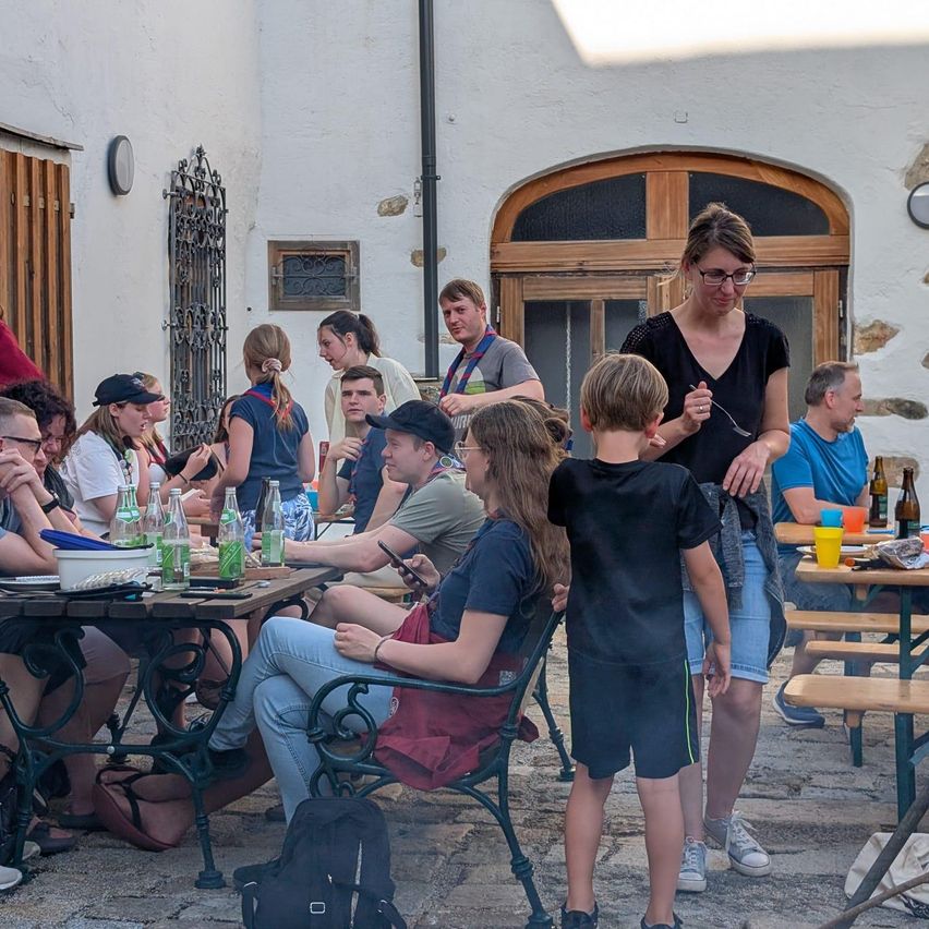 Eine Gruppe von Menschen sitzt draußen an Tischen. Flaschen, Becher und Teller sind auf den Tischen. Eine Frau mit Brille steht und hält einen Löffel. Hinter ihr steht ein Junge und hält ein Handy. Eine Frau sitzt auf einem Stuhl.