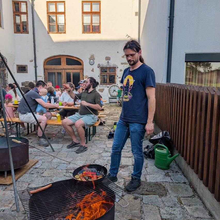 Ein Mann grillt draußen auf einem Grill, während Menschen an Picknicktischen sitzen. Er trägt ein blaues T-Shirt mit Grafikdesign. In der Nähe steht ein grüner Gießkanne auf dem Boden.