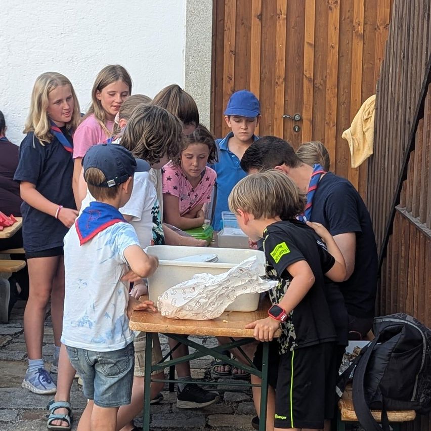 Eine Gruppe von Kindern und Jugendlichen versammelt sich um einen Tisch mit Essen darauf. Einige tragen Abzeichen und Mützen. Hinter ihnen ist eine Holztür zu sehen.