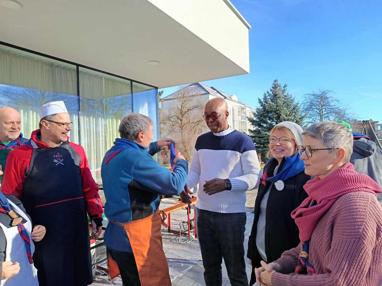 A group of people are standing outdoors. A man in a red jacket is grilling. Another man in a blue jacket is holding a medal.