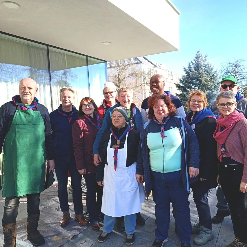 A group of people are posing for a photograph in front of a building with glass walls. Some are wearing glasses, scarves, and boots. They are standing in a row, smiling. Behind them, there is a tree and a building.
