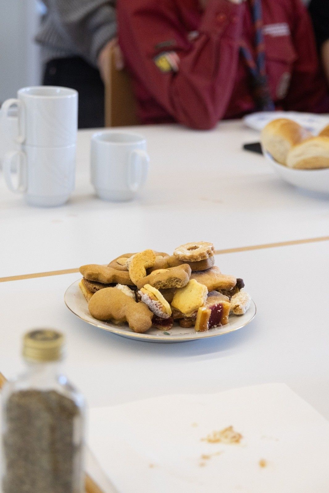Ein weißer Tisch zeigt einen Teller mit verschiedenen Keksen, eine Schüssel mit Brot und zwei weiße Tassen. Eine Flasche mit goldener Kappe steht links.