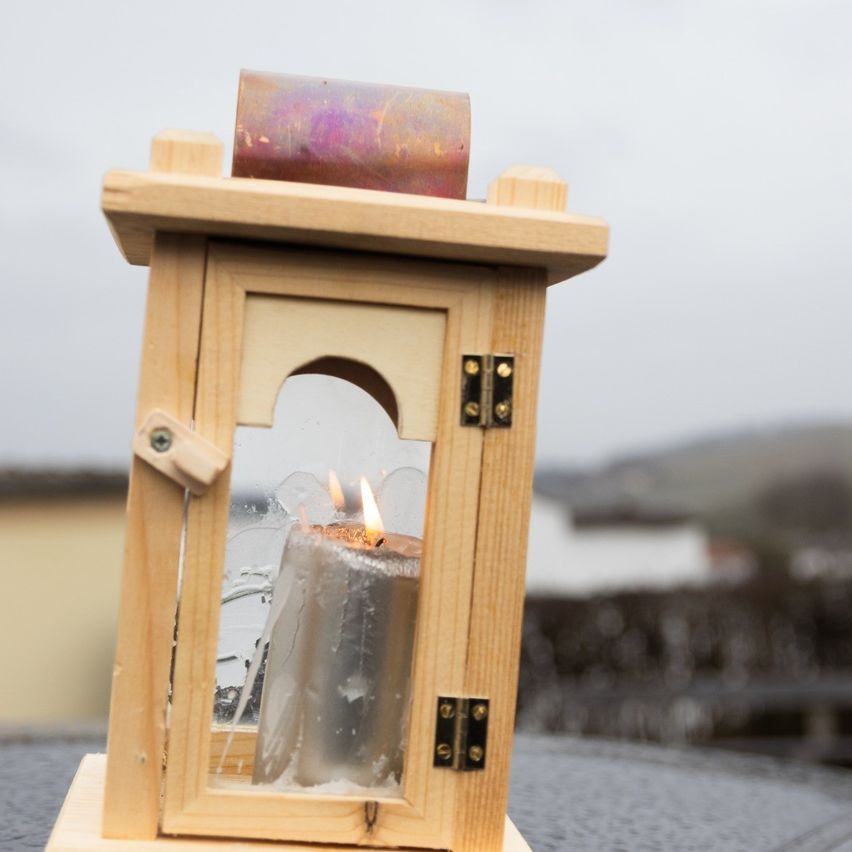 Eine hölzerne Laterne mit einer brennenden Kerze darin steht auf einem Tisch. Die Laterne hat ein Glasfenster und einen Metalldeckel oben. Die Umgebung ist verschwommen.
