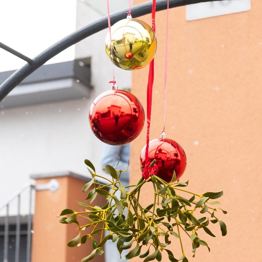 Eine Reihe roter und goldener Weihnachtskugeln hängen an einem roten Band, das an einem Gebäude befestigt ist. Die Kugeln reflektieren ihre Umgebung.