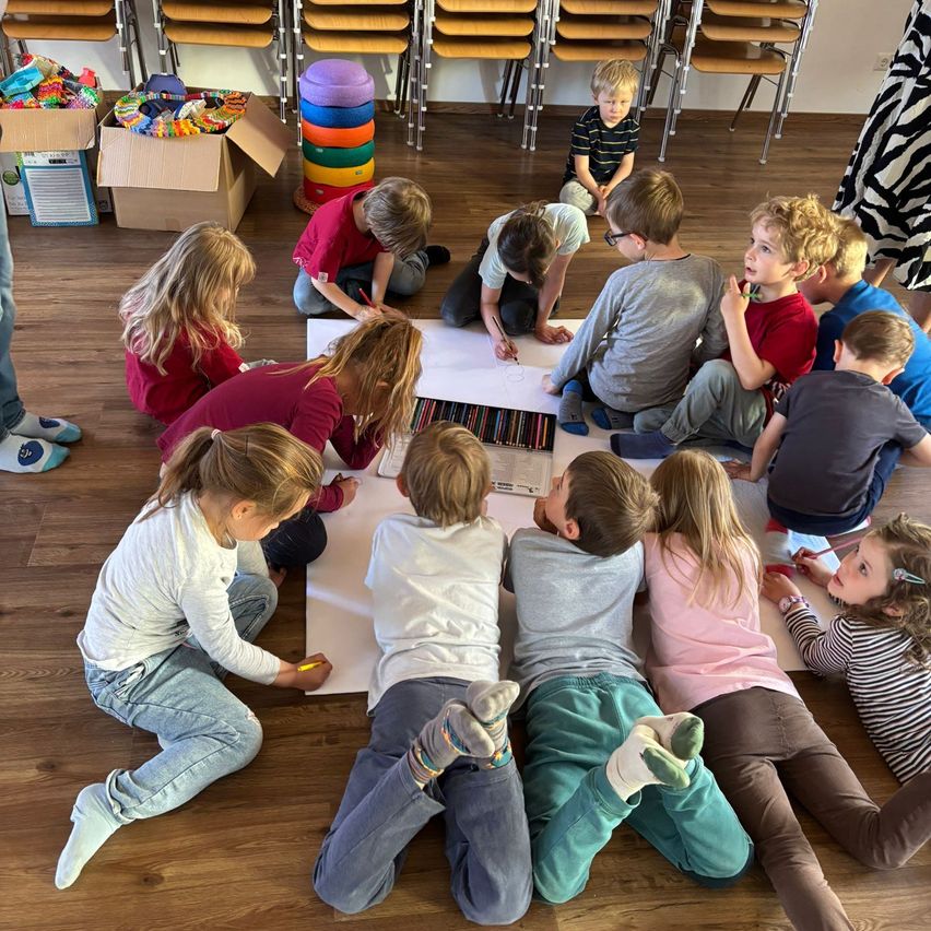 Eine Gruppe von Kindern sitzt im Klassenzimmer auf dem Boden, zeichnet und färbt mit Buntstiften auf einem großen Stück Papier. Einige tragen eine Brille.