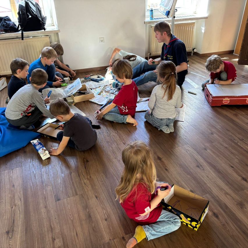 Eine Gruppe von Kindern sitzt auf dem Boden in einem Raum und beschäftigt sich mit verschiedenen Aktivitäten. Einige zeichnen, während andere Bücher lesen. Ein Mann steht in der Nähe und beobachtet sie.