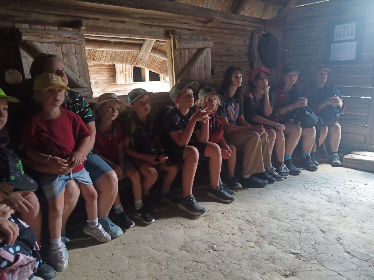 Eine Gruppe von Kindern und Erwachsenen sitzt auf einer Bank in einer Holzhütte. Sie tragen alle Mützen und Turnschuhe.