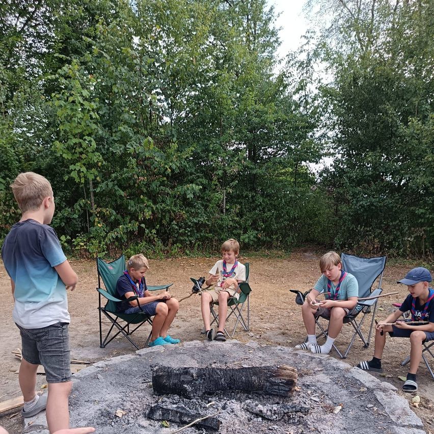 Fünf Jungen sitzen in einem Wald um ein Lagerfeuer und tragen Pfadfinderuniformen. Ein Junge hält einen Marshmallow am Stiel.