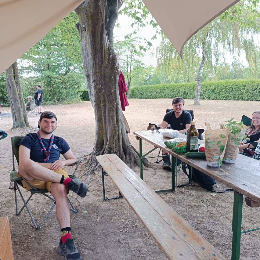 Eine Gruppe von Menschen sitzt um einen Picknicktisch in einem Park, ein Mann trägt ein Pfadfinderabzeichen und ein anderer hat eine Flasche Wein auf dem Tisch.