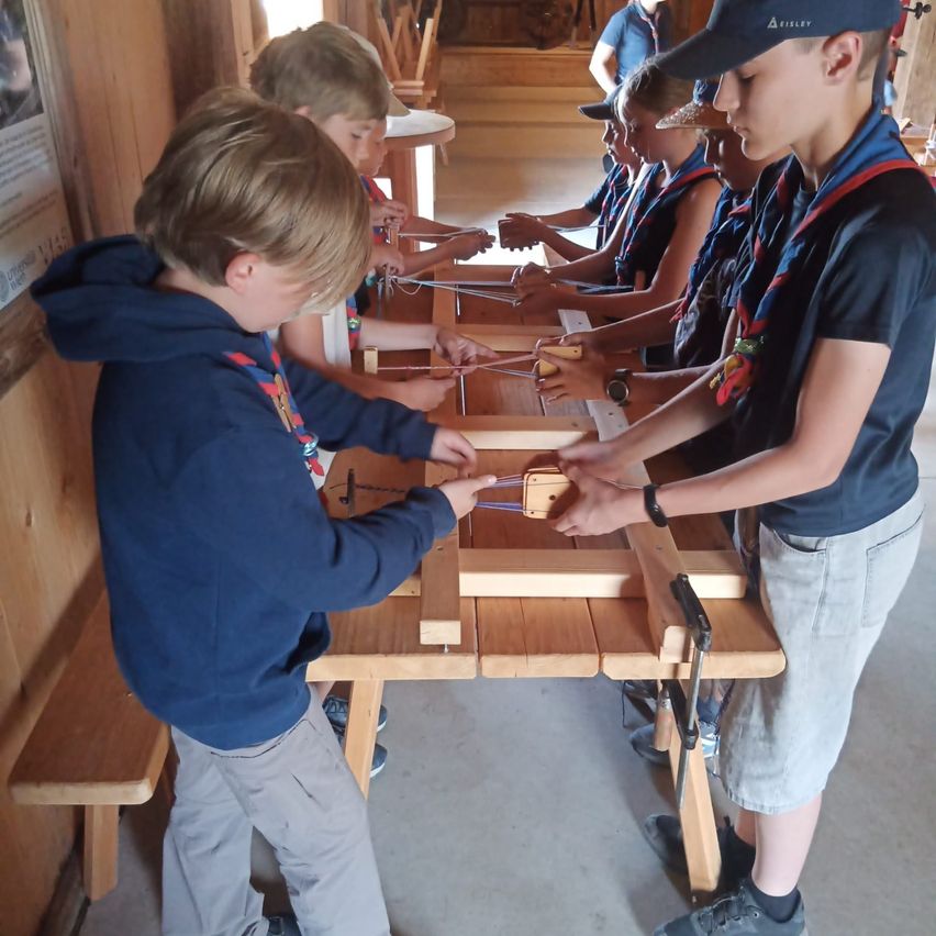 Eine Gruppe junger Jungen in Pfadfinderuniformen arbeitet in einer Werkstatt an einer Holzstruktur, wobei ein Junge ein Werkzeug benutzt, um einen Teil zu justieren.