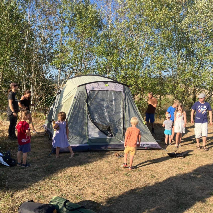Eine Gruppe von Menschen, einschließlich Kinder, versammelt sich um ein großes Zelt in einem Wald. Einige stehen, während andere in der Nähe des Zeltes sind. Das Zelt ist teilweise geöffnet, und der Bereich ist von Bäumen umgeben.
