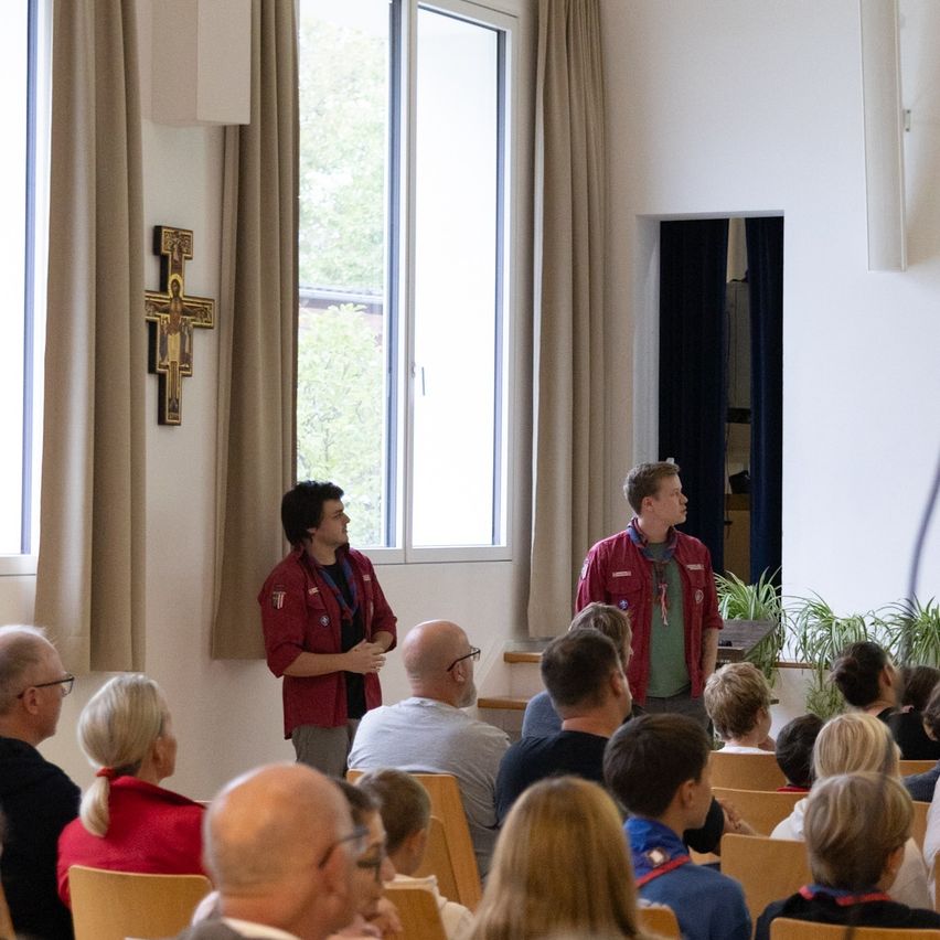 Zwei Männer stehen vor einem Raum voller Menschen. Ein Mann trägt eine rote Jacke und der andere ein grünes Hemd.