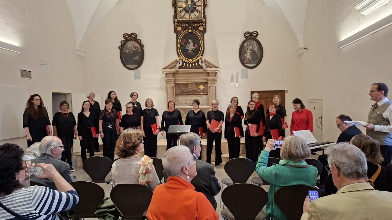 Ein Chor in schwarzen Kleidern hält rote Ordner und steht vor einem Altar, eine Uhr und Gemälde schmücken die Wand. Einige Zuschauer beobachten vom Stuhl aus.
