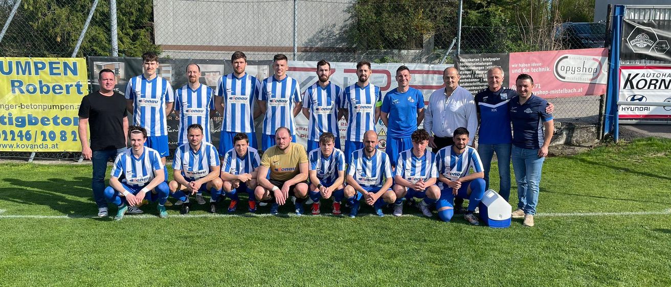 Eine Fußballmannschaft in blau-weißen Trikots posiert für ein Mannschaftsfoto auf einem grünen Feld mit einem Gebäude und Bäumen im Hintergrund.