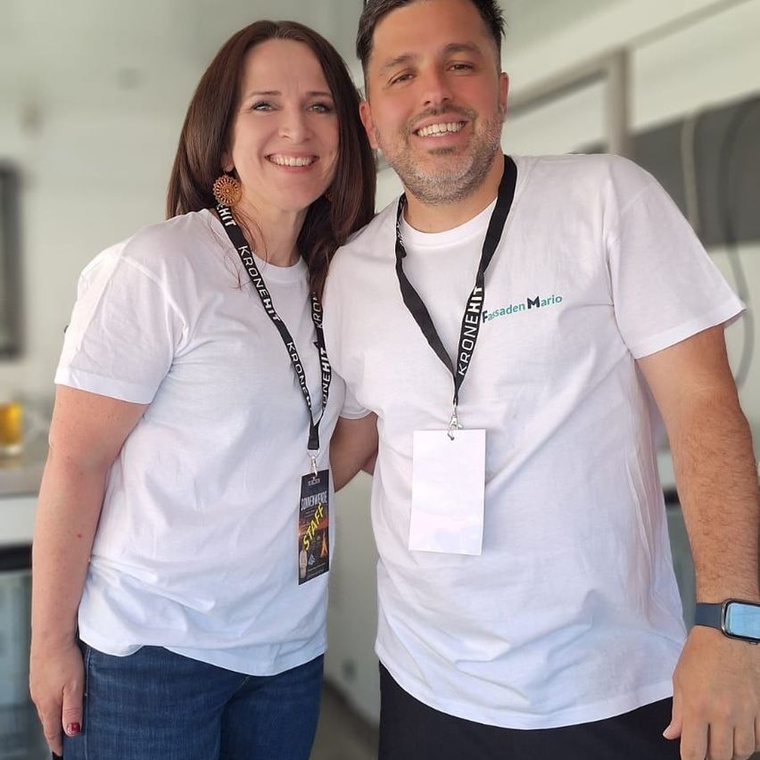 Ein lächelndes Paar in weißen T-Shirts und Lanyards mit 'Kronenhit'-Logo und 'Pasadena Mario'-Text posiert für ein Foto.