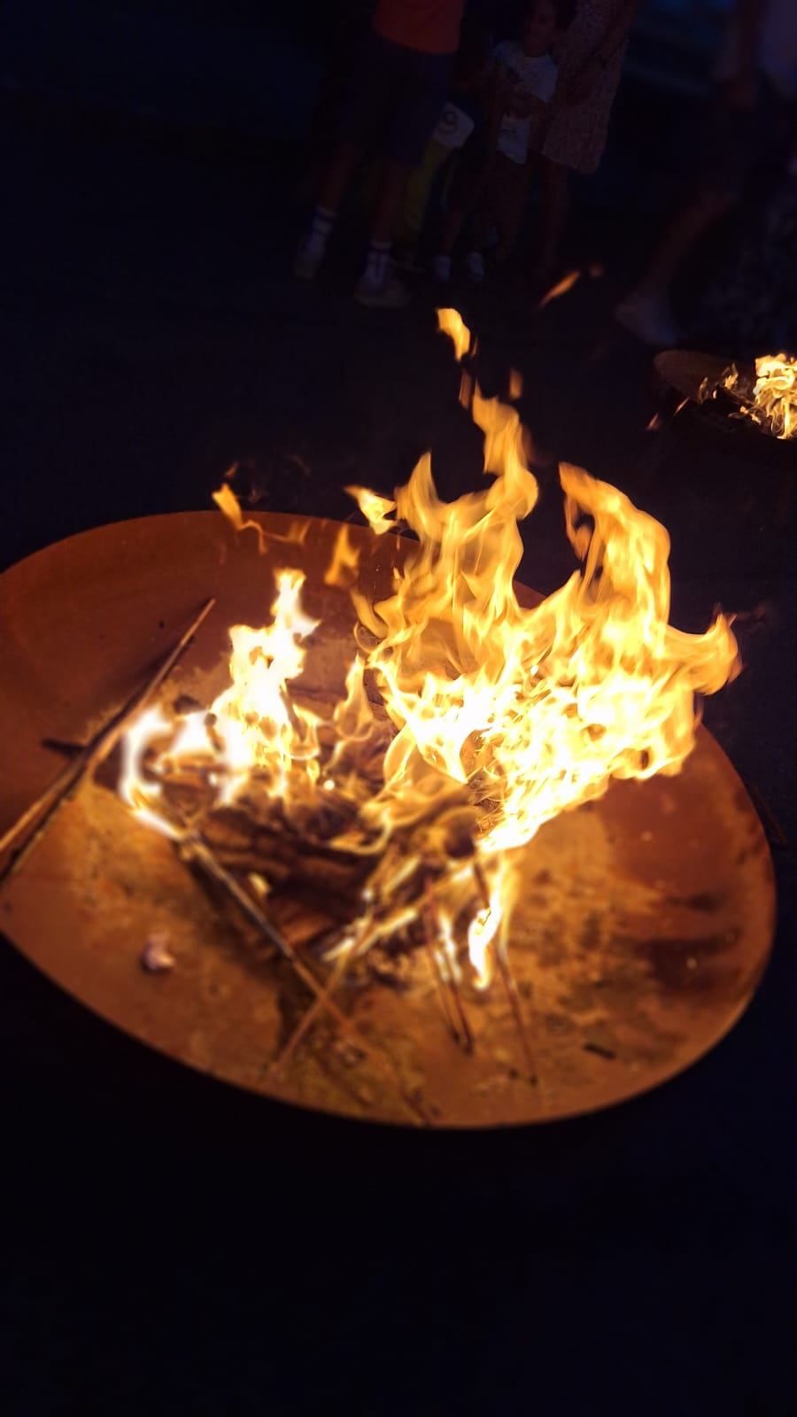 Ein Lagerfeuer brennt hell in einer Metallschale und wirft warmes Licht und flackernde Flammen gegen die Dunkelheit.