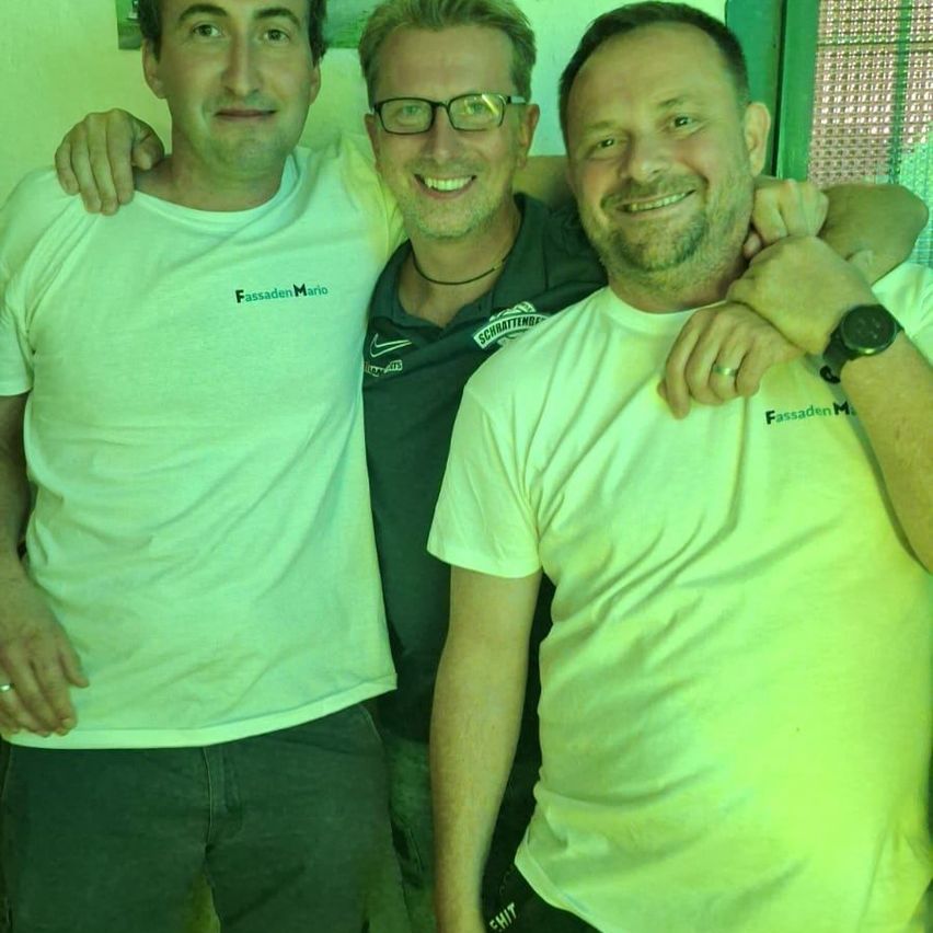 Drei Männer posieren für ein Foto, alle tragen helle T-Shirts mit Text. Der Mann links trägt ein weißes Shirt mit dem Wort 'FassadenMario' darauf.