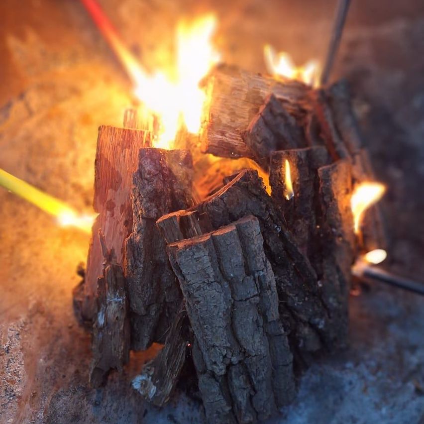 Ein Holzstapel brennt mit hellen Flammen auf einer sandigen Oberfläche. Das Feuer strahlt ein warmes Licht aus.