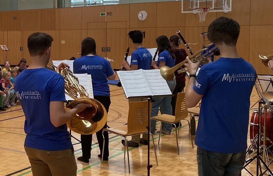 Eine Gruppe von Menschen in blauen Shirts spielt Musikinstrumente in einem Raum. Eine Uhr ist an der Wand montiert. Ein Basketballkorb ist an der Wand rechts angebracht.
