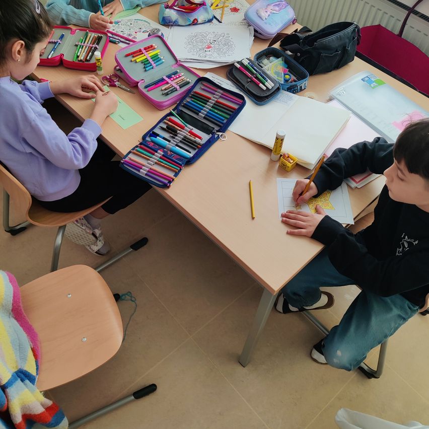 Kinder sitzen um einen Tisch herum und beschäftigen sich mit Malaktivitäten. Ein Kind malt auf Papier, während ein anderes Malwerkzeuge auf dem Tisch verteilt hat.