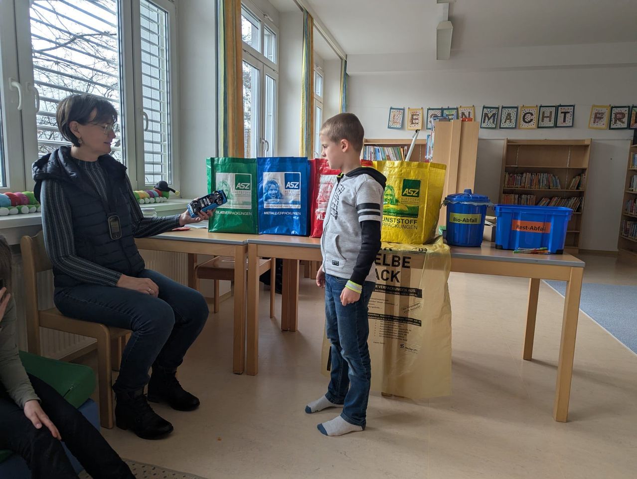 Eine Frau hält ein Handy und spricht mit einem Jungen. Sie befinden sich in einem Klassenzimmer mit Recyclingbehältern, einem Schreibtisch und einem Bücherregal.