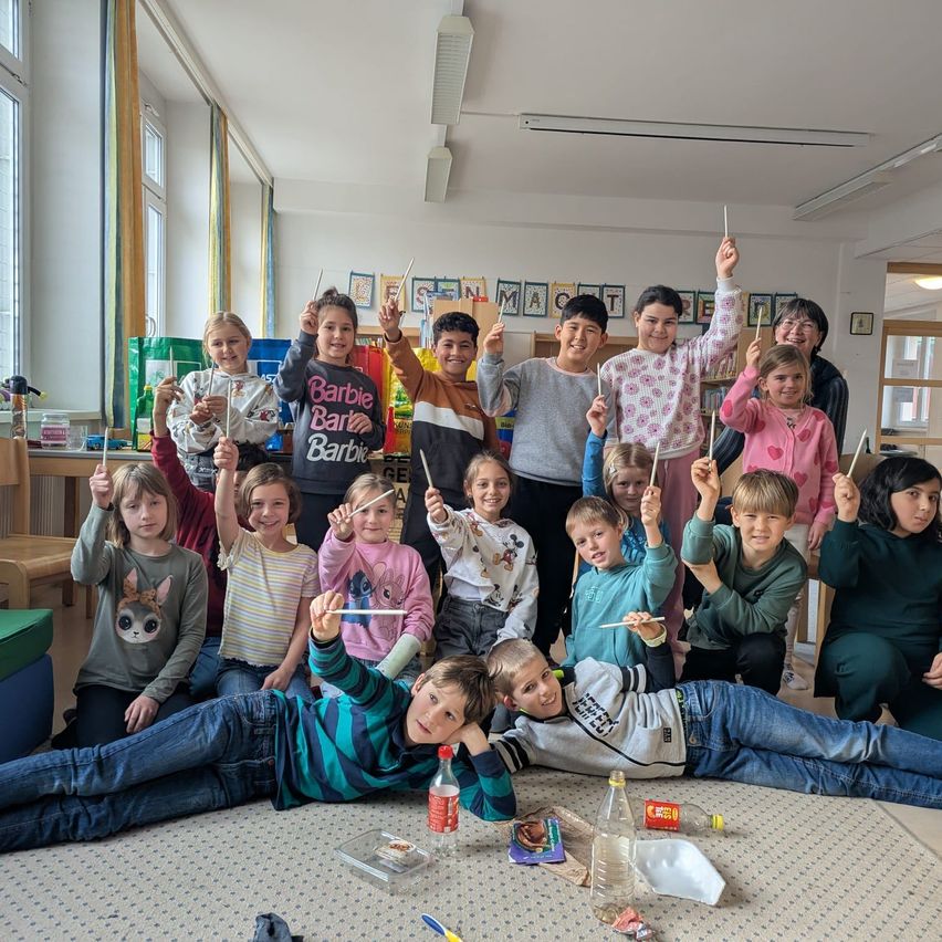 Eine Gruppe von Kindern in einem Klassenzimmer hält Stöcke, einer liegt auf dem Boden, mit einem Tisch, Stühlen, Flaschen und einer Tür im Hintergrund.