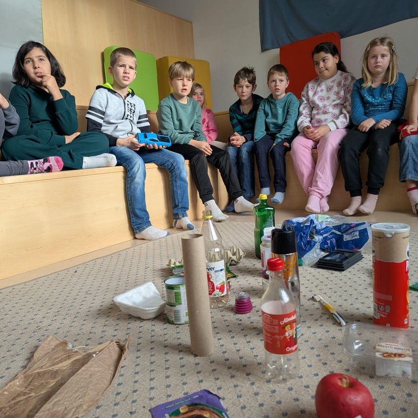 Eine Gruppe von Kindern sitzt auf Bänken in einem Klassenzimmer und sieht der Kamera entgegen. Sie wirken nachdenklich. Verschiedene Gegenstände wie eine Flasche, Dosen und ein Papierbeutel sind auf dem Boden verstreut. Ein Apfel liegt ebenfalls auf dem Boden.