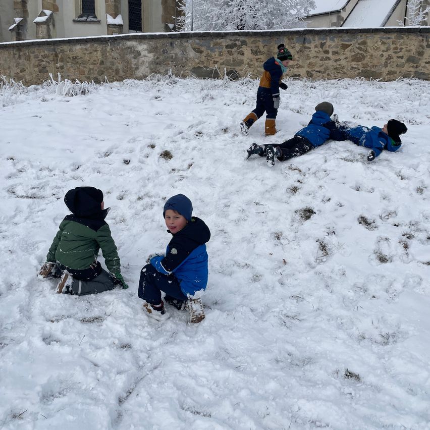Kinder spielen im Schnee vor einer Steinmauer. Ein Kind sitzt, während andere auf dem Schnee liegen.