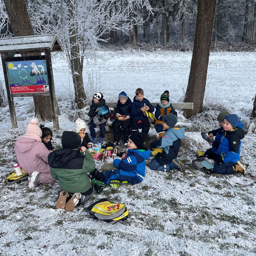 Eine Gruppe von Kindern sitzt im Schnee in einem Wald und trägt Winterkleidung. Einige essen. Hinter ihnen steht ein Holzschild.