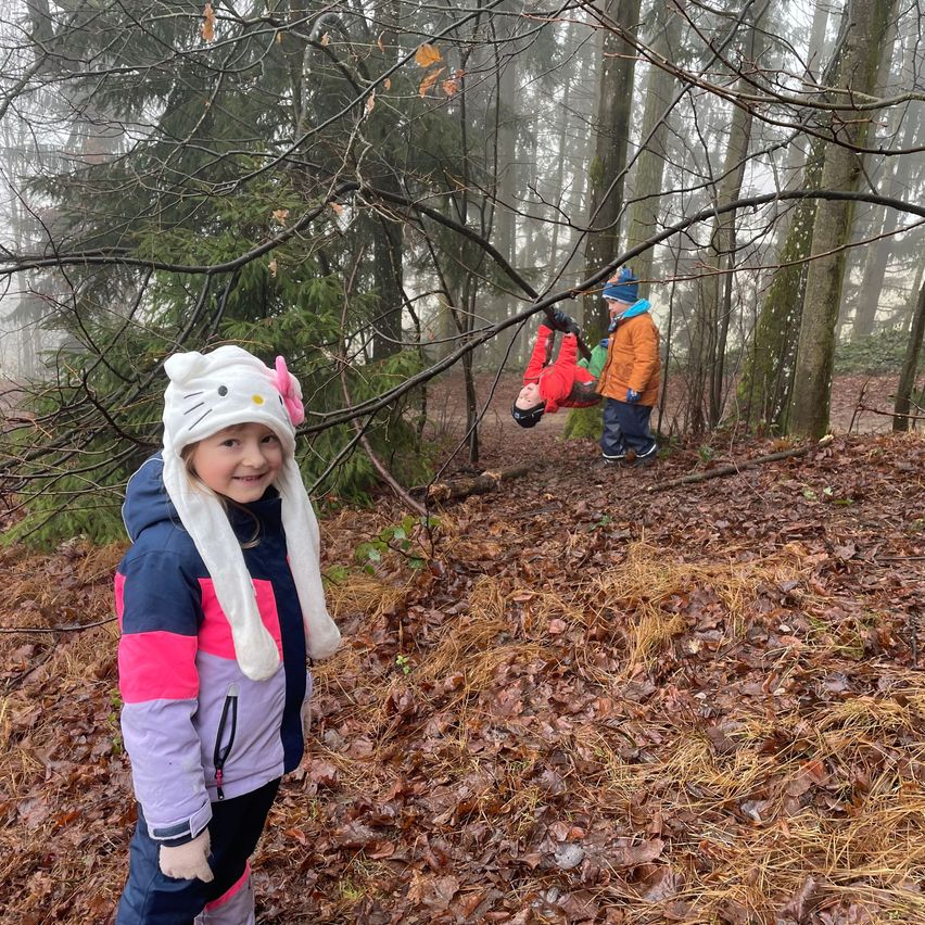Ein junges Mädchen in einer bunten Jacke steht in einem Wald, während ein Junge an einem Ast über ihr schwingt. Der Wald ist neblig und der Boden ist mit gefallenen Blättern bedeckt.