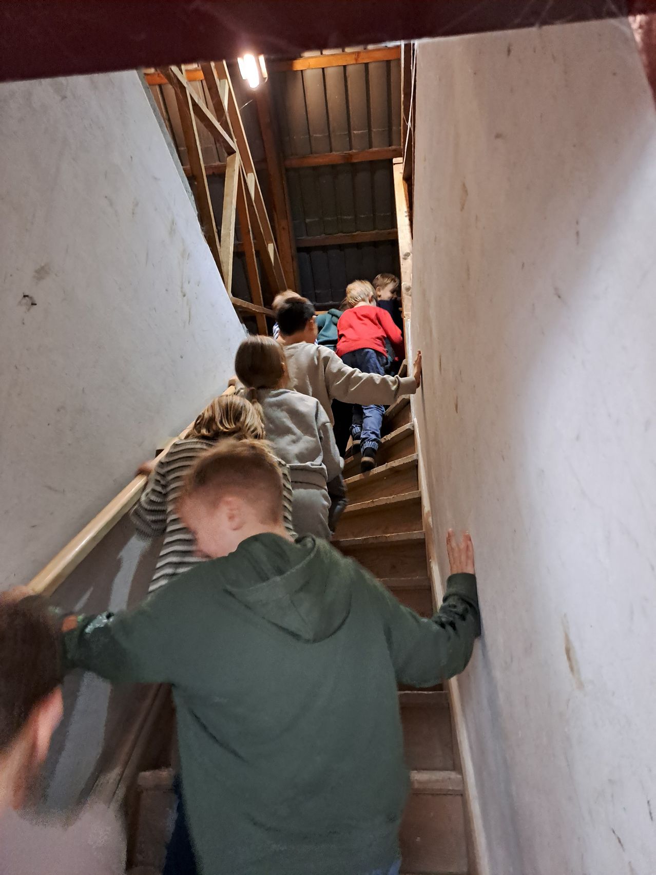 Eine Gruppe von Kindern steigt eine Holztreppe hinauf. Die Wände sind weiß gestrichen. Die Kinder halten sich an den Geländern fest.
