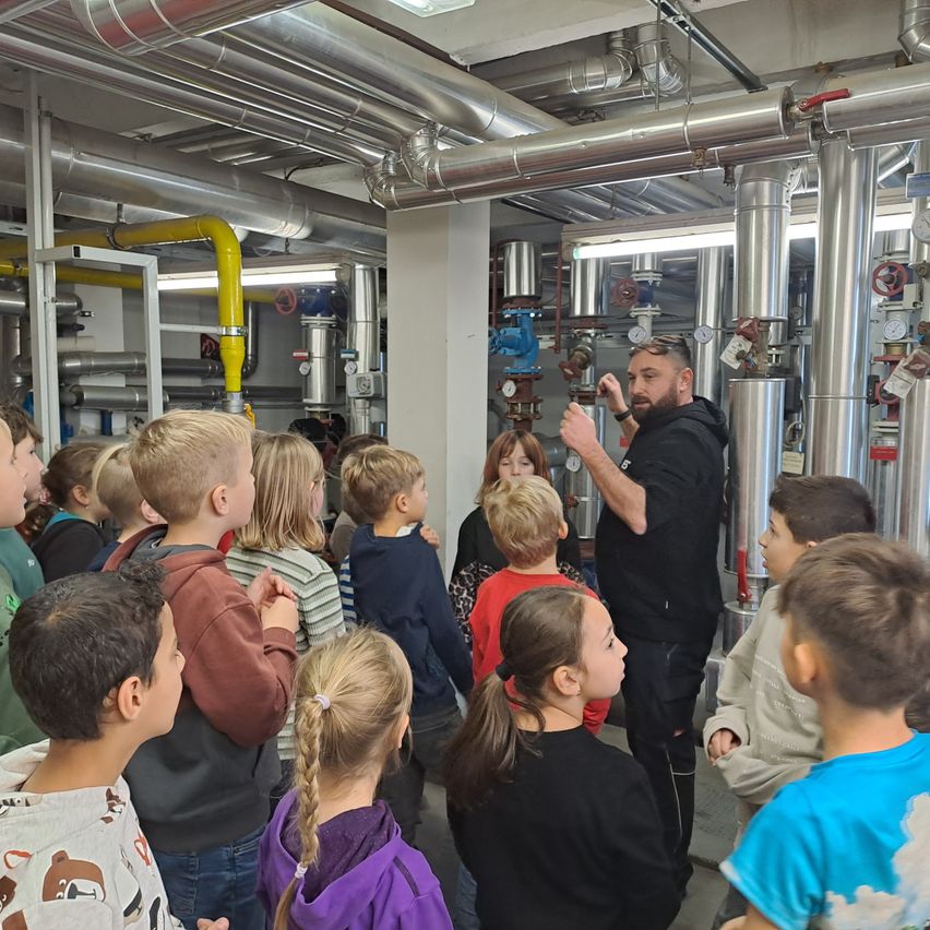 Ein Mann in einer schwarzen Kapuzenjacke erklärt etwas einer Gruppe von Kindern in einem Raum mit Metallrohren und Maschinen.