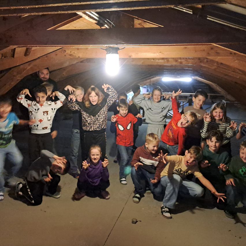 Eine Gruppe von Kindern und Erwachsenen posiert für ein Foto in einem hölzernen Dachboden mit einer hängenden Lampe. Die Kinder tragen verschiedene thematische Shirts, und einige machen Handgesten.