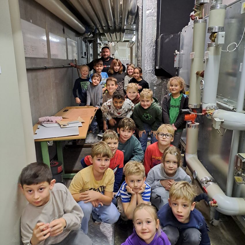 Eine Gruppe von Kindern und Erwachsenen versammelt sich in einem Korridor und posiert für ein Foto. Einige knien, andere stehen. Im Hintergrund sind Rohre und Maschinen zu sehen.