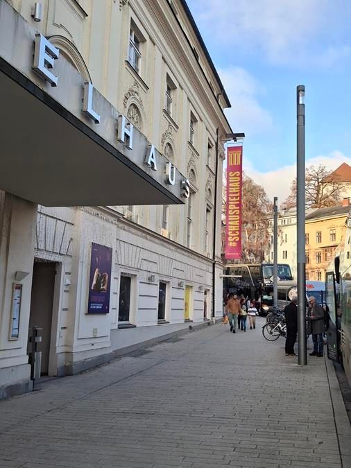 Ein Blick auf die Straße des Elhaus-Gebäudes, eines Theaters mit einem Banner zur Förderung von #schauspielhaus. Menschen gehen vorbei und ein Bus ist in der Nähe geparkt.