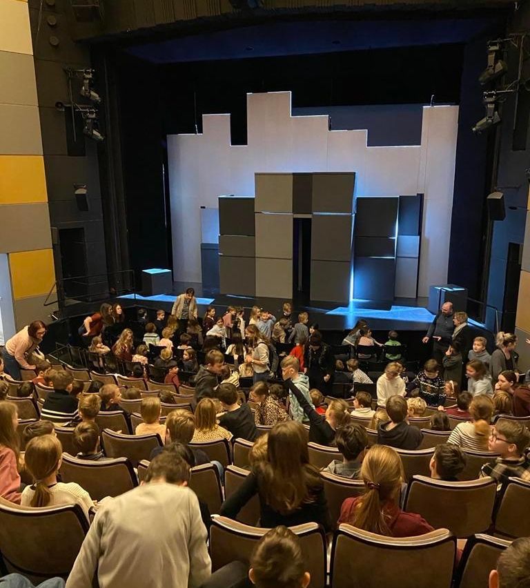 Eine große Menge an Kindern und Erwachsenen sitzt in einem Theater und schaut sich eine Bühnenaufführung mit Publikum an.