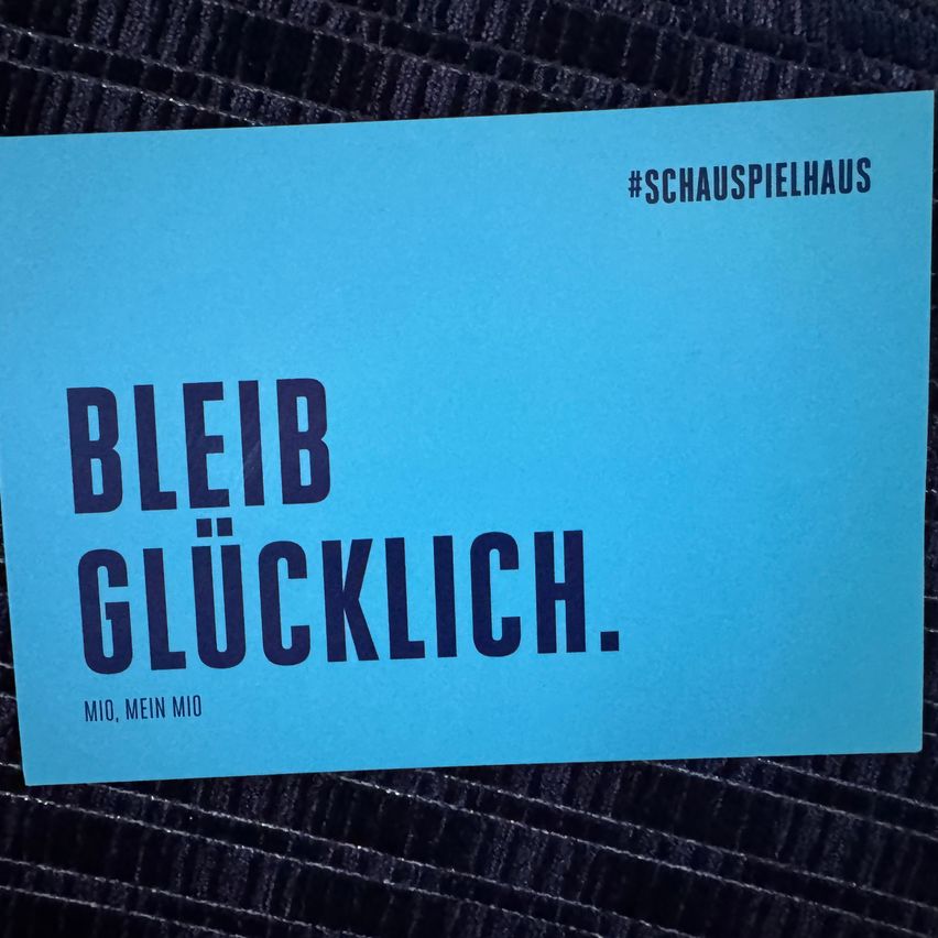 Eine blaue Karte zeigt in großen schwarzen Buchstaben 'BLEIB GLÜCKLICH'. Darunter steht der Slogan '#SCHAUSPIELHAUS'. In kleineren Buchstaben steht 'MIO, MEIN MIO'.