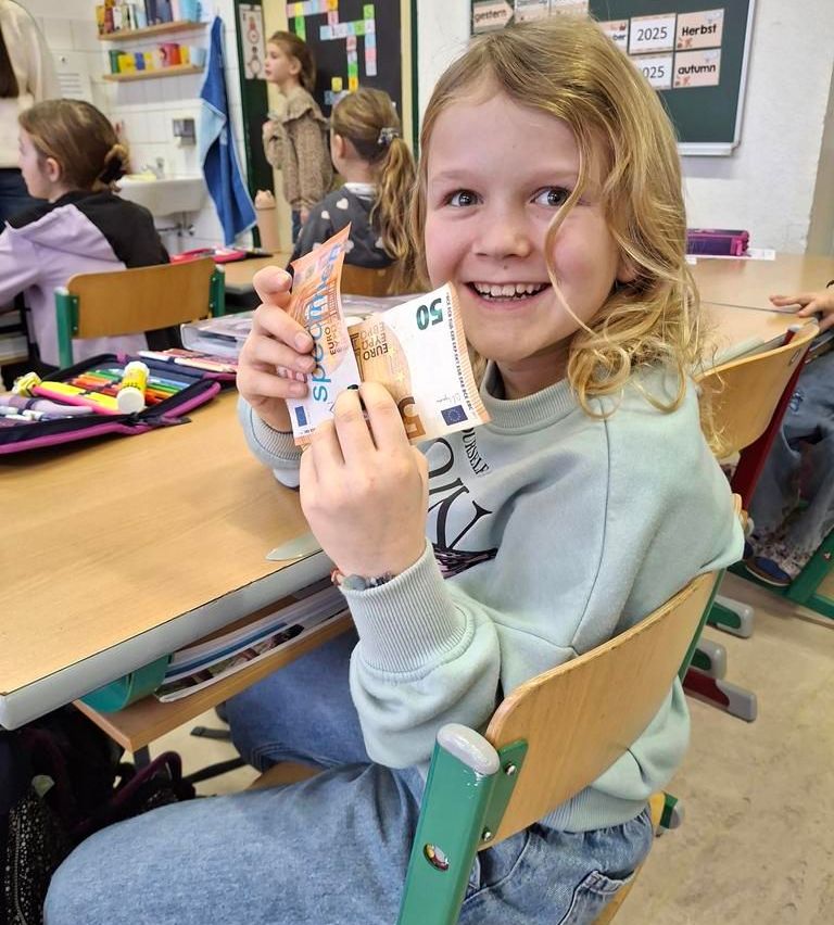 Ein kleines Mädchen im Klassenzimmer hält eine Euro-Banknote und lächelt, umgeben von Schulsachen und anderen Kindern.