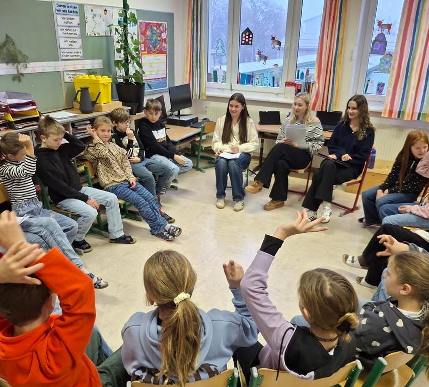 Ein Klassenzimmer mit Kindern, die im Kreis sitzen, zwei Frauen mit Papieren in der Mitte. Einige Schüler heben die Hände. Ein Heizkörper und Fenster mit Dekorationen sind im Hintergrund.