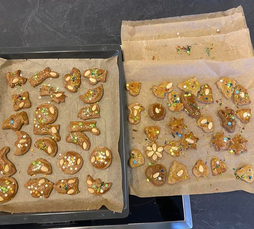 Zwei Backbleche gefüllt mit dekorierten Keksen, einige mit Mandeln darauf und andere mit bunten Streuseln. Die Bleche stehen auf einer grauen Oberfläche.