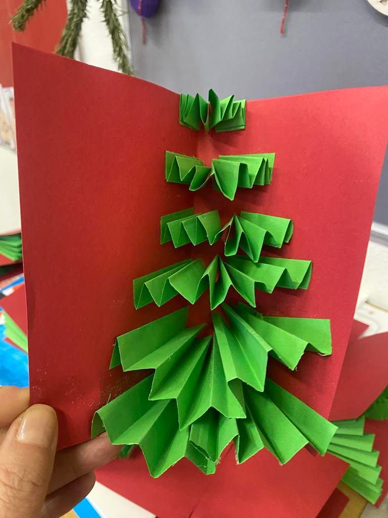 Ein Nahaufnahme einer handgefertigten Weihnachtsbaumkarte aus grünem Papier, auf rotem Hintergrund platziert. Die Karte zeigt kompliziertes Falten, das einen 3D-Effekt erzeugt.