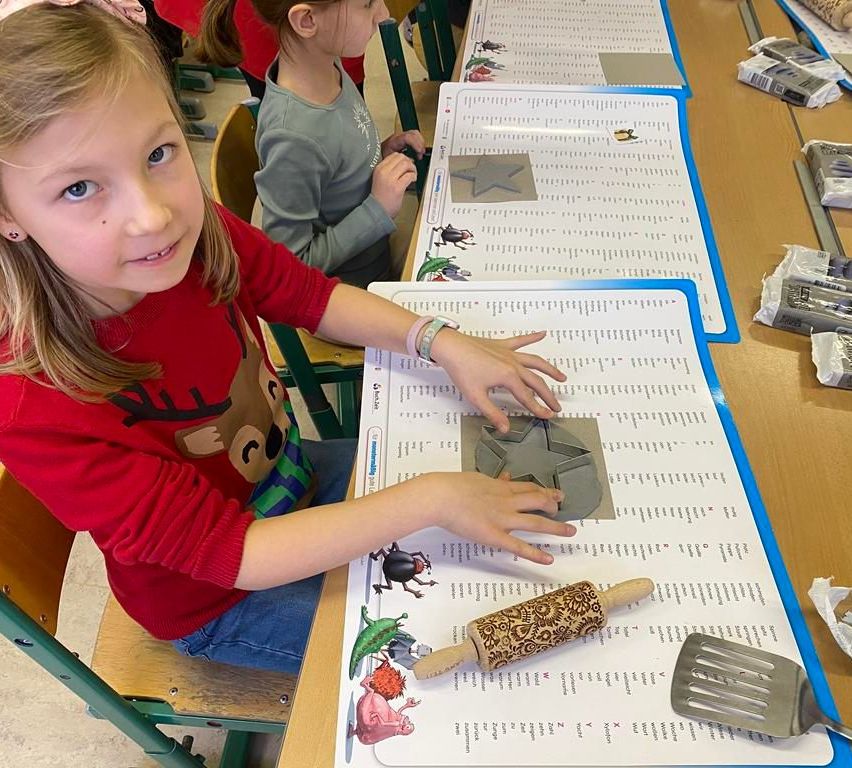 Ein kleines Mädchen in einem roten Shirt benutzt einen hölzernen Roller, um ein sternförmiges Objekt auf einem Tisch zu kreieren, der mit einem weißen Blatt Papier bedeckt ist.