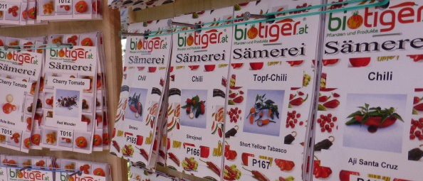 An einer Wand sind mehrere Samentüten für verschiedene Pflanzen zu sehen, darunter Kirschtomaten und Chilis, mit eindeutigen Produktcodes und Bildern auf jedem Beutel.
