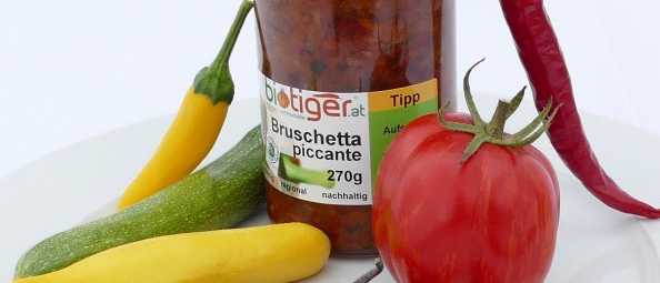 Auf einem weißen Teller sind verschiedene Gemüsesorten und ein Glas Bruschetta Piccante zu sehen. Dazu gehören Karotten, Zucchini, gelbe Zucchini und Auberginen. Ein roter Tomaten und zwei Paprikaschoten, eine rot und eine grün, sind ebenfalls vorhanden.