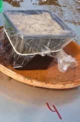 Ein durchsichtiger Plastikbehälter mit einer weißen Substanz ist mit Plastikfolie versiegelt auf einem braunen Teller. Der Teller liegt auf einer weißen Oberfläche mit einer roten Zahl vier.